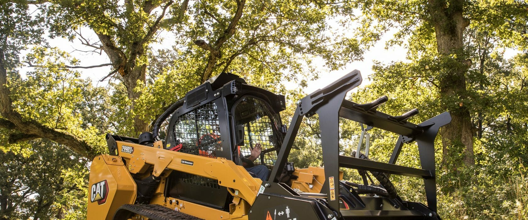 cat machine in the middle of the forest with a large attachment on the front and the sun coming through the trees